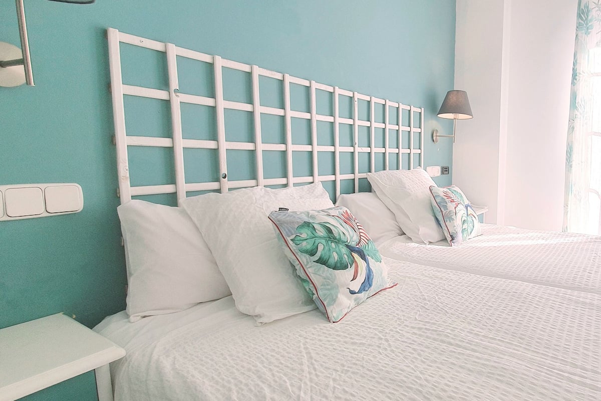 Une chambre d’hôtel avec deux lits, une tête de lit blanche, une table de chevet, une lampe et une fenêtre avec des rideaux.