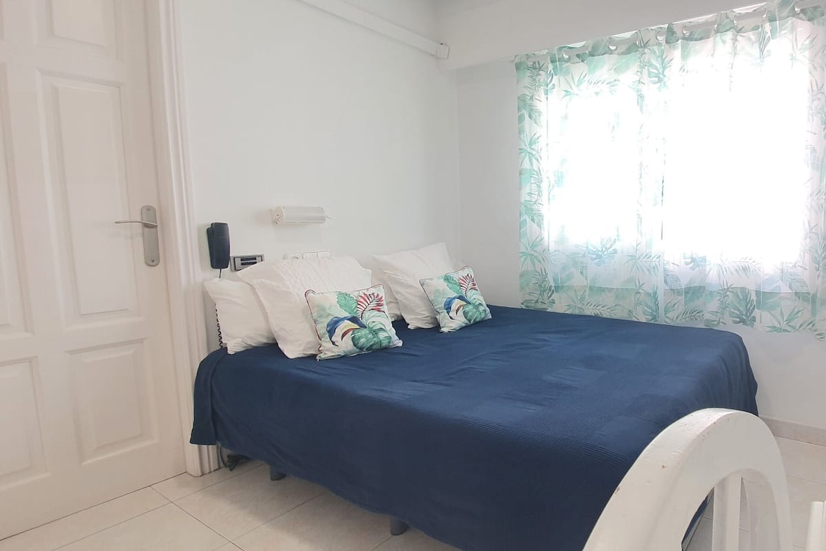 Une chambre à coucher avec un lit, des oreillers, une fenêtre ornée d’un rideau à motifs floraux et une porte blanche.