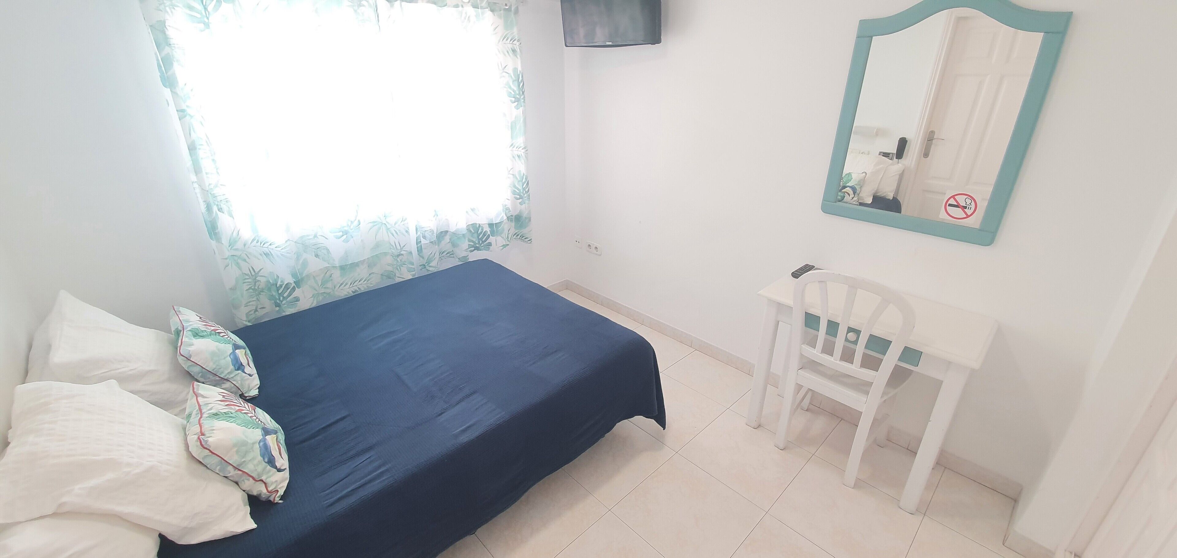 Une chambre à coucher comprenant un lit, une chaise, une fenêtre avec un rideau, un miroir et un téléviseur fixé au mur.