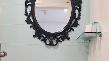 Une salle de bain avec un lavabo blanc, un miroir rond doté d’un cadre décoratif, une étagère en verre et un tabouret blanc.