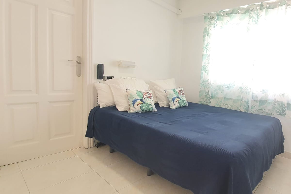 Une chambre à coucher avec un lit, des oreillers, une table de chevet, une fenêtre avec un rideau à motifs floraux et une porte blanche.