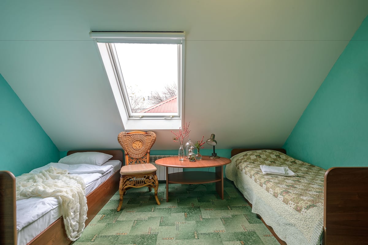 A small, triangular room with two beds, a chair, a small table, and a window.