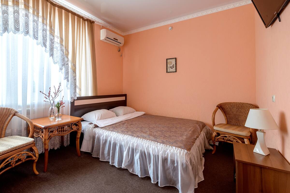 A hotel room with a single bed, a bedside table with a lamp, a wicker chair, a small table with a vase, and an air conditioning unit on the wall.