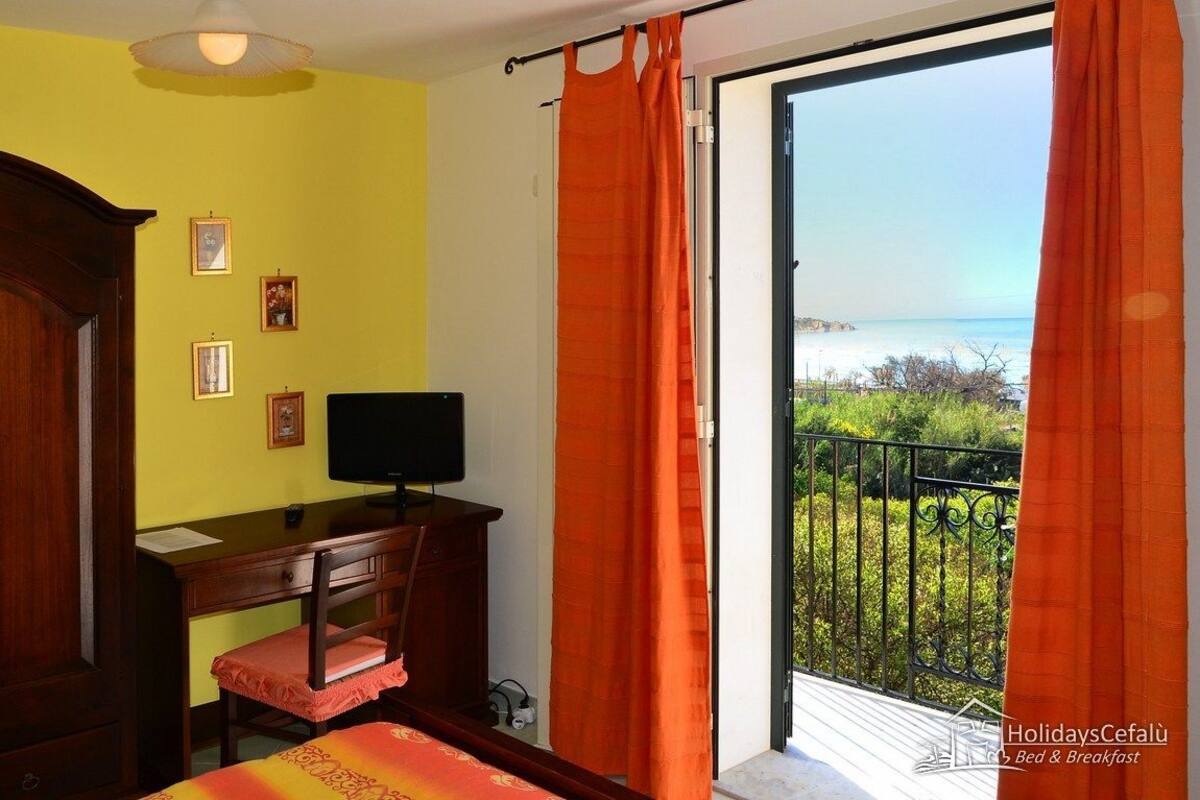 Una habitación con un escritorio de madera, una silla, una cama con colcha roja y amarilla, un televisor sobre el escritorio, y un balcón con cortinas naranjas y vistas al mar.