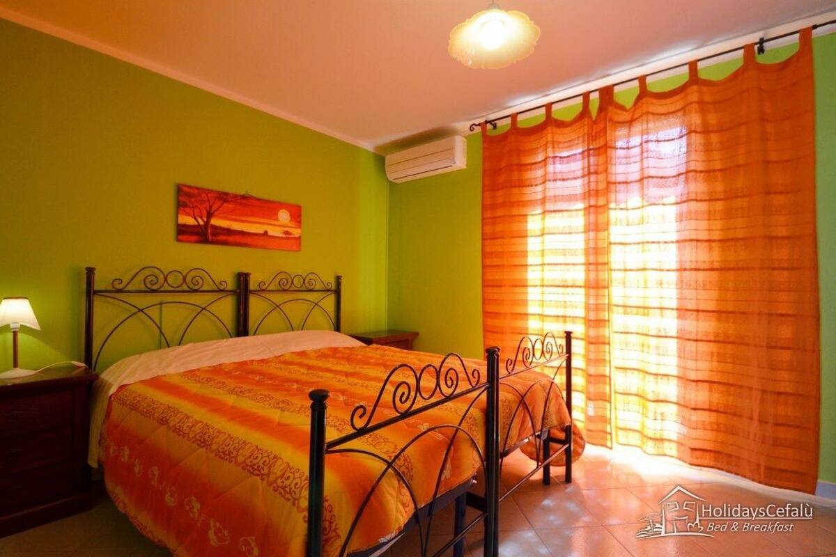 Un dormitorio con cama, mesita de noche, lámpara, un cuadro en la pared y ventana con cortinas.