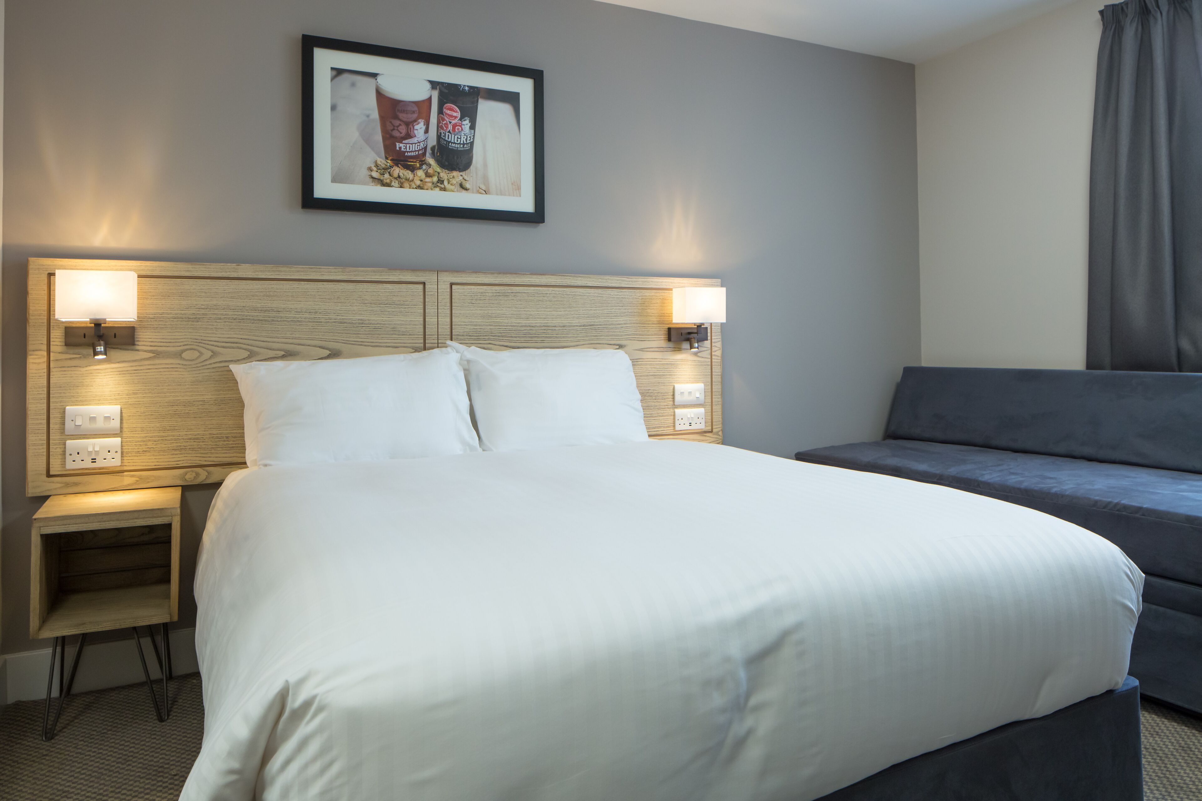Habitación de hotel con una cama grande, lámparas de noche, un sofá gris y un cuadro enmarcado en la pared.