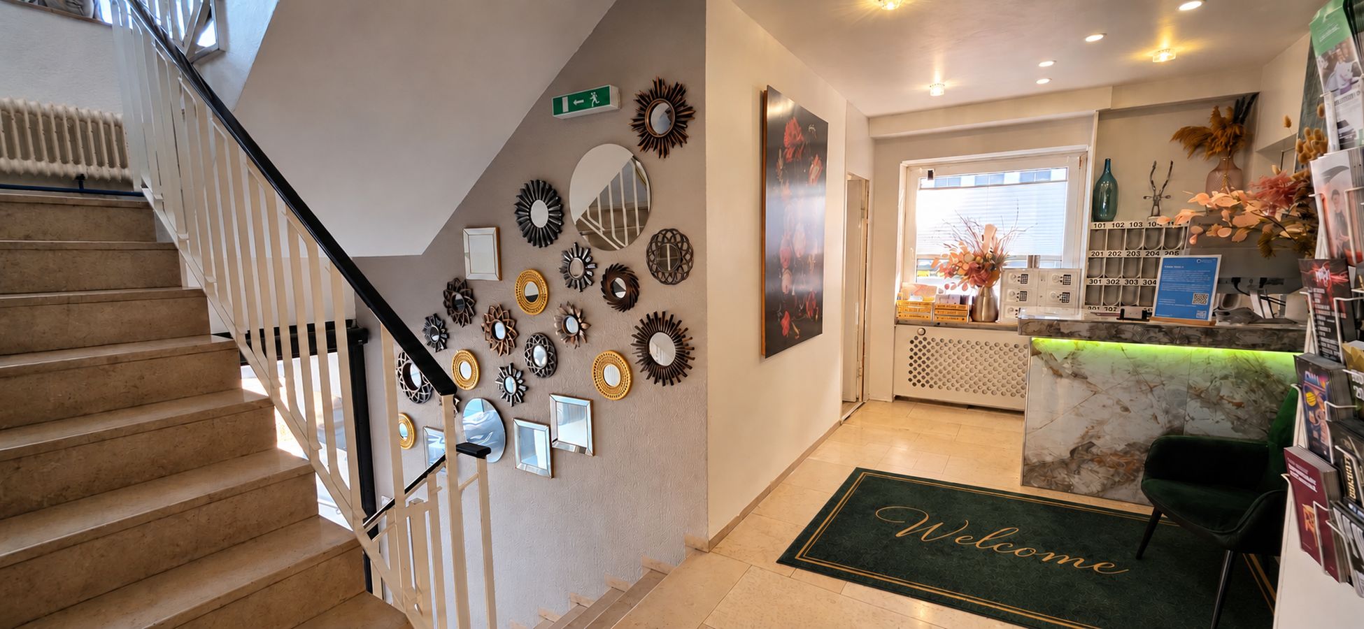 A hotel lobby with a staircase, a reception desk, and a wall decorated with various mirrors.