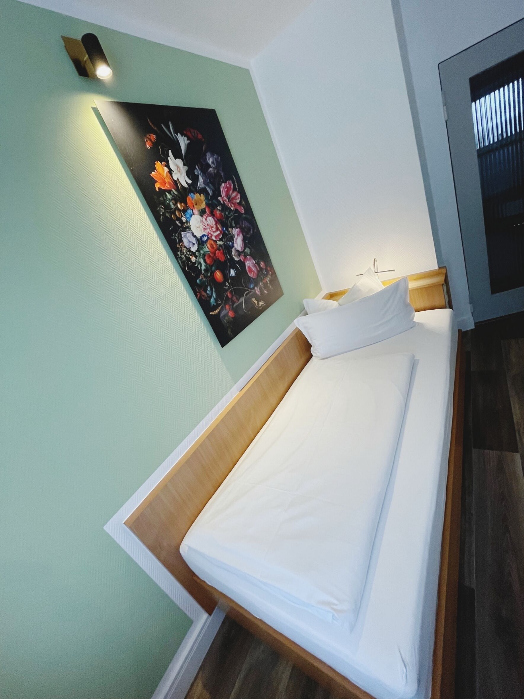 Una cama bien tendida en una habitación con pared verde y un cuadro floral sobre ella.