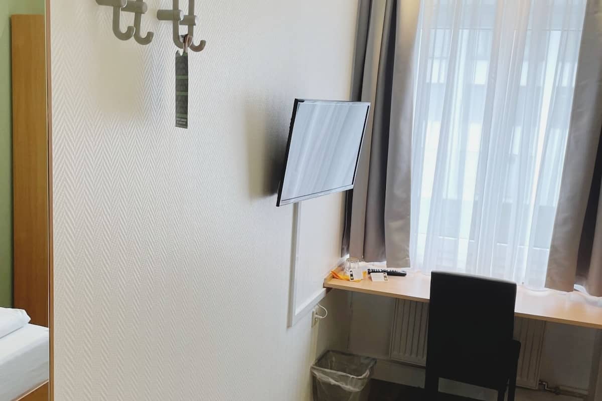 Ett hotellrum med en säng, ett skrivbord med en dator och en bildskärm, en stol, ett fönster med gardiner och en papperskorg.