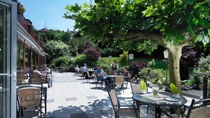 Un espace extérieur aménagé en terrasse, avec des tables et des chaises, entouré de verdure et bordé d’un bâtiment doté de grandes fenêtres.