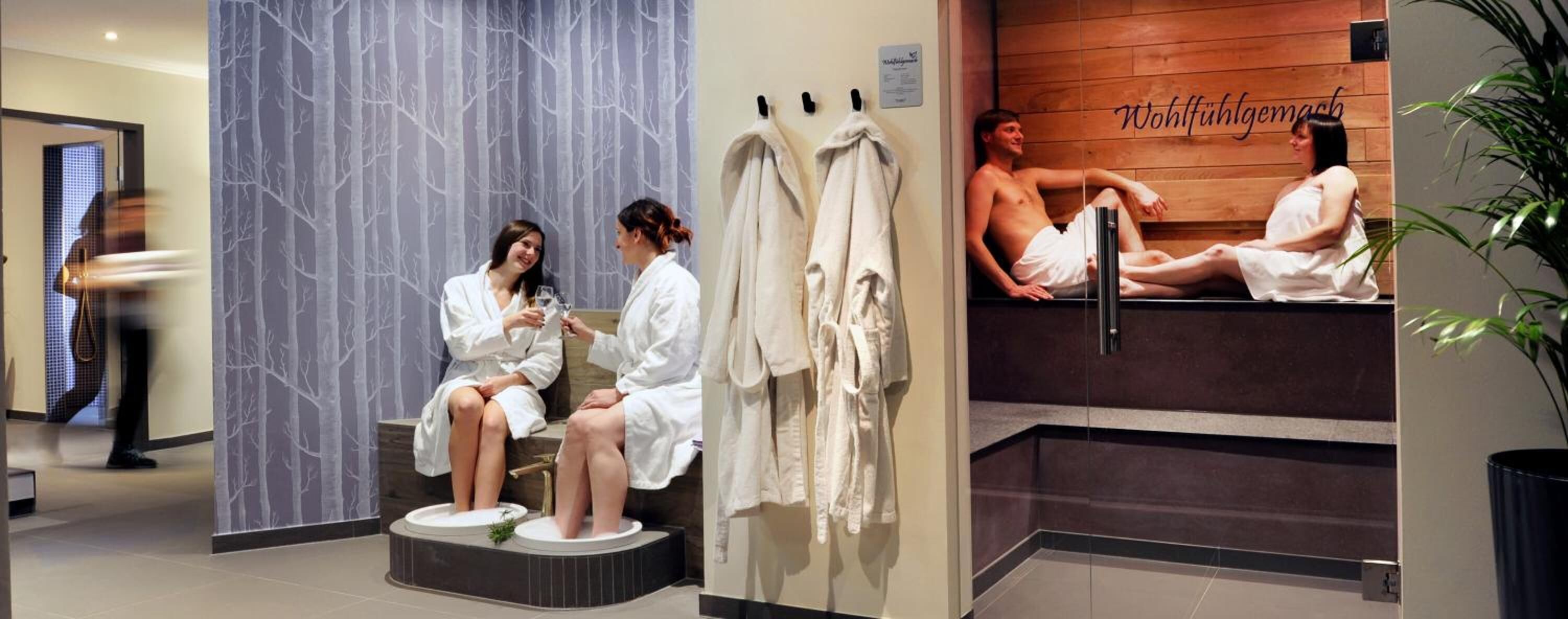 A spa area with two people in robes, a wooden wall with a towel rack, and a poster of a man and woman in a bath.