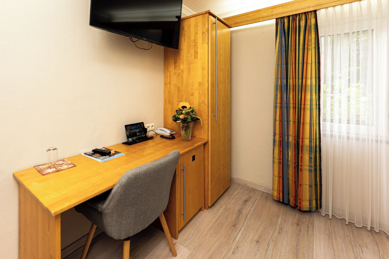 Une pièce de taille modeste, avec un bureau en bois, une chaise grise, une petite plante et un téléviseur fixé au mur.