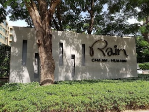 Placa indicando 'Rain Cha Am - Huahin' com uma árvore e vegetação em primeiro plano.