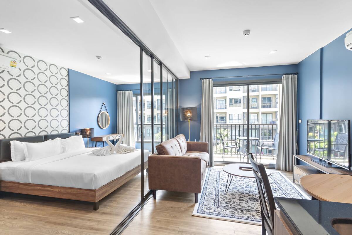A modern hotel room with a large bed, a sofa, a TV, and a balcony with a view of the city.