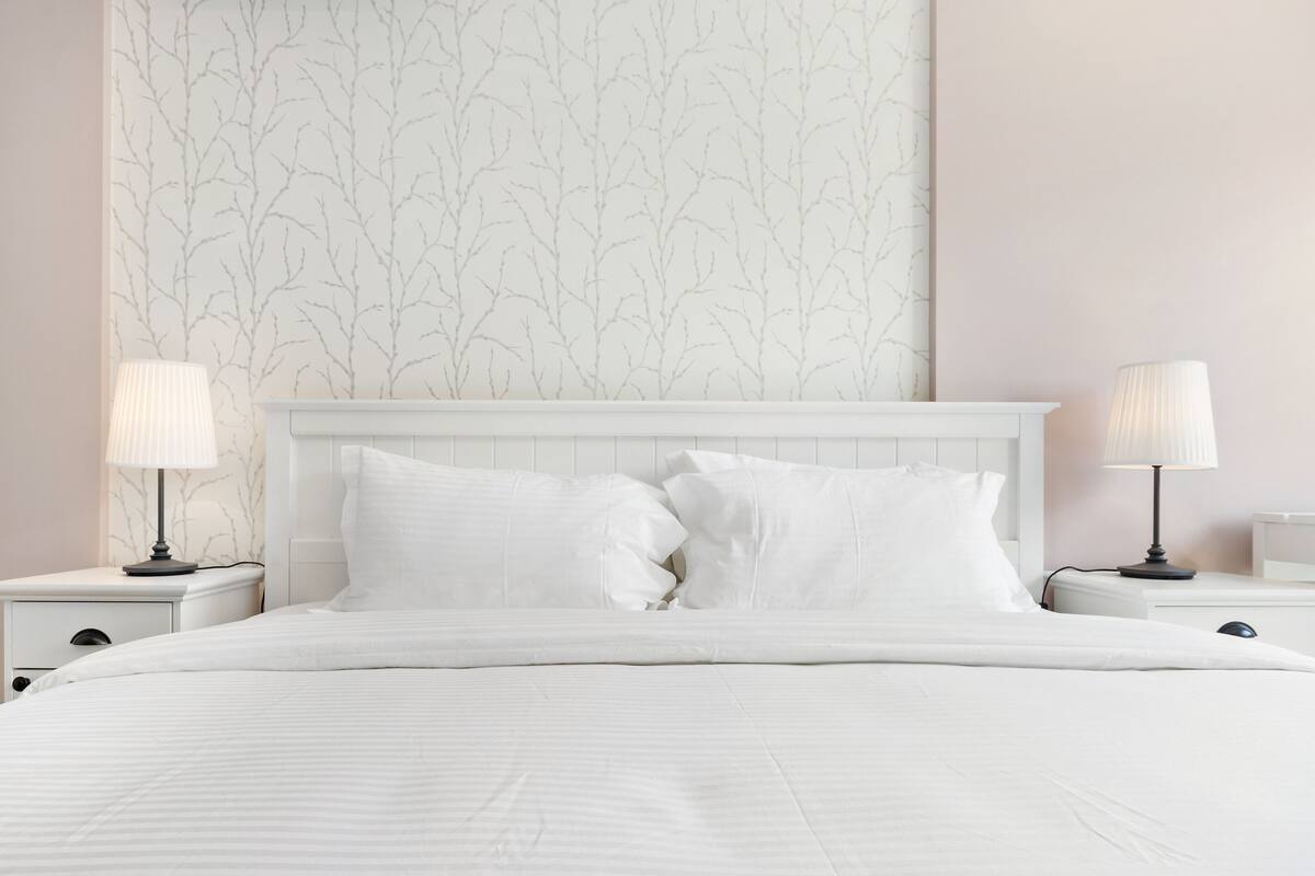 A neatly made bed with white linens and two bedside lamps, set against a wall with a subtle tree pattern.