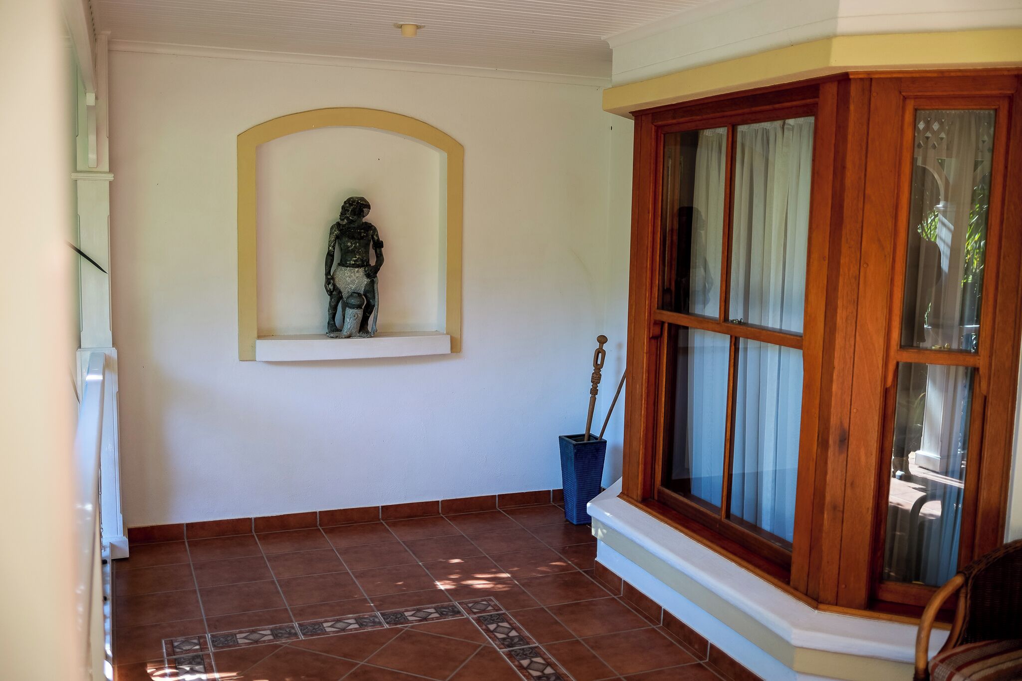 A room with a tiled floor, a statue on a shelf, a window with curtains, and a wooden cabinet.