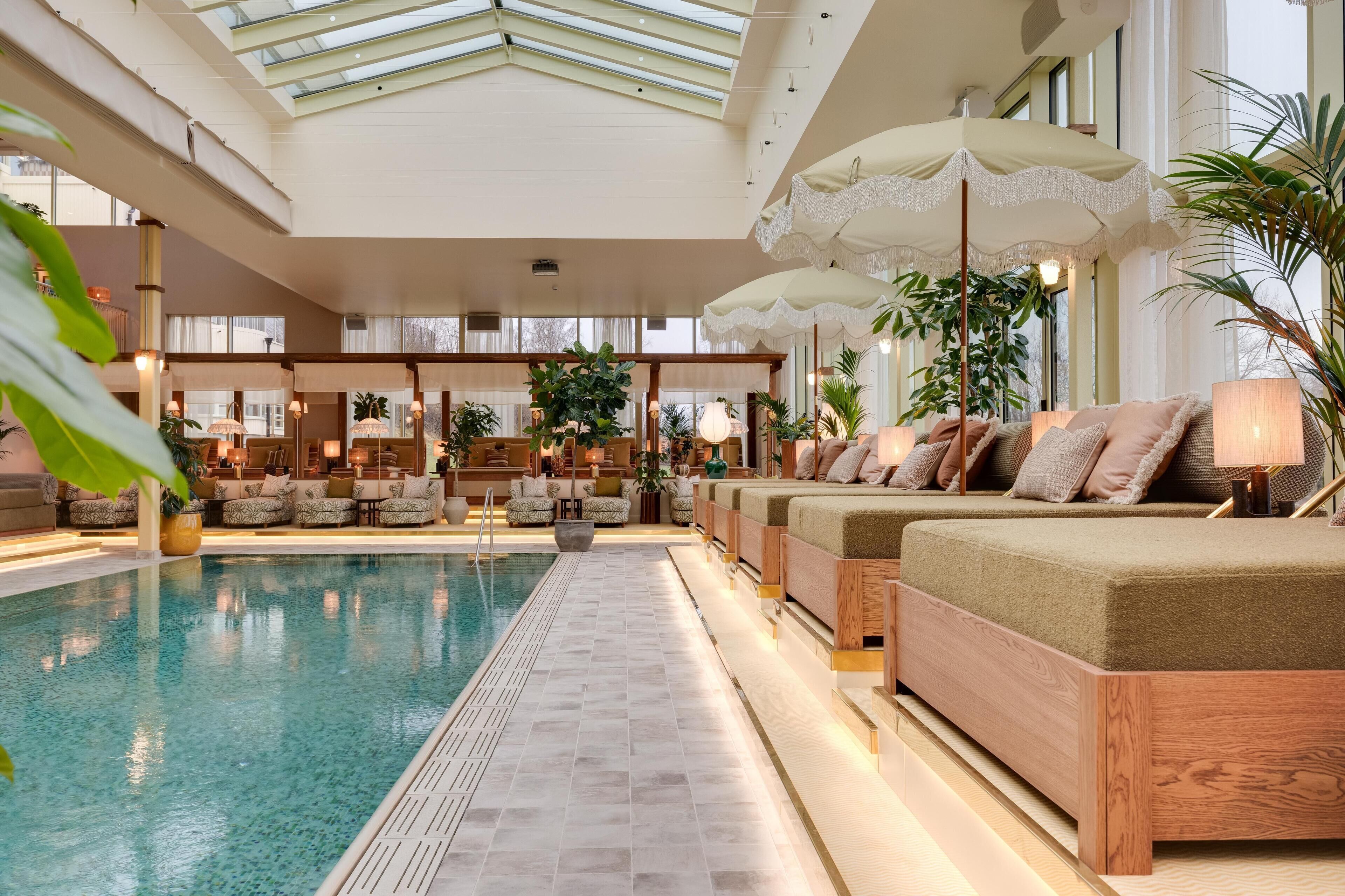 A spacious indoor pool area with a long bench seating arrangement, surrounded by various plants and lamps.