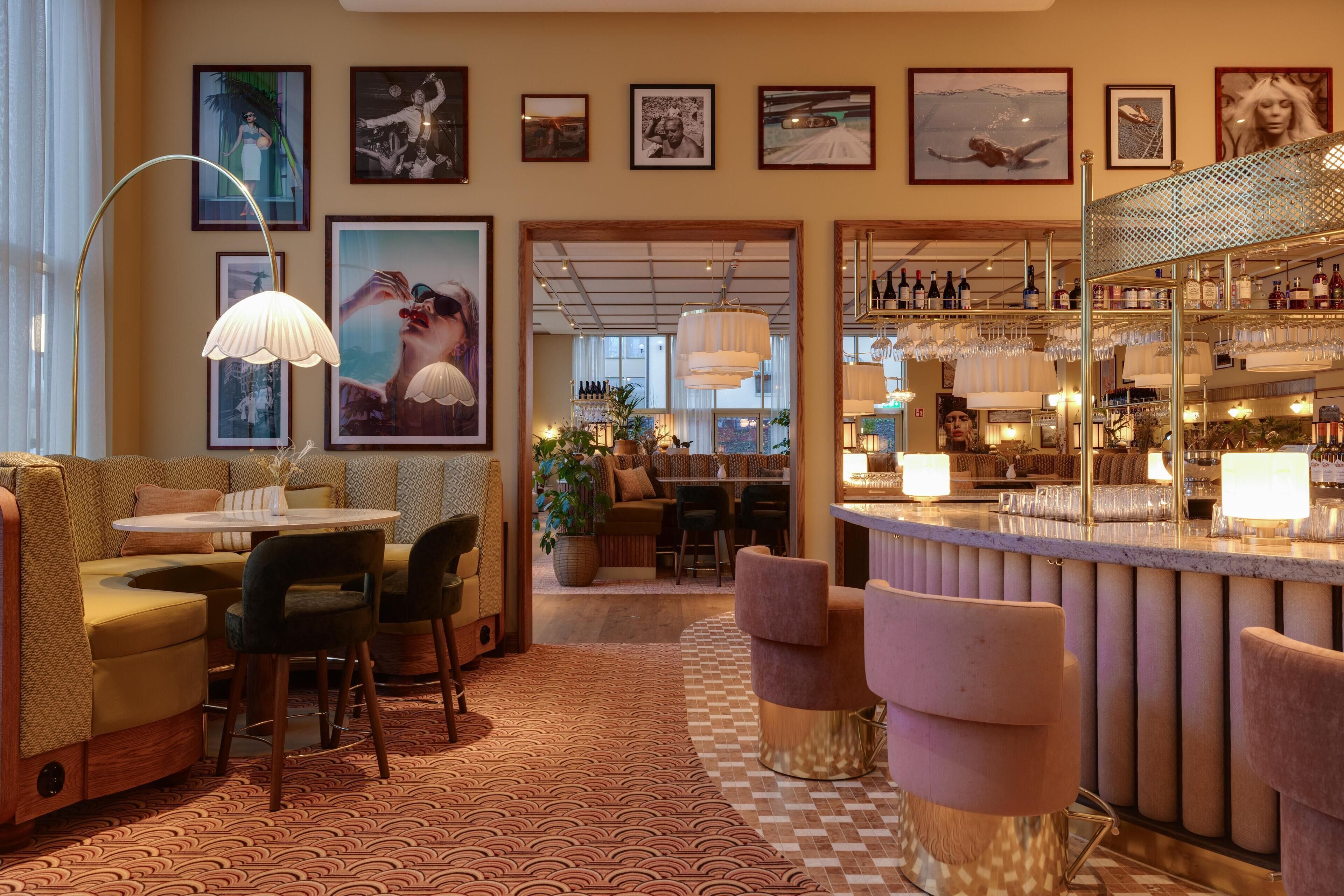 Een bar met een zwart-wit geruit vloertje, barstoelen en een toog vol flessen en glazen.