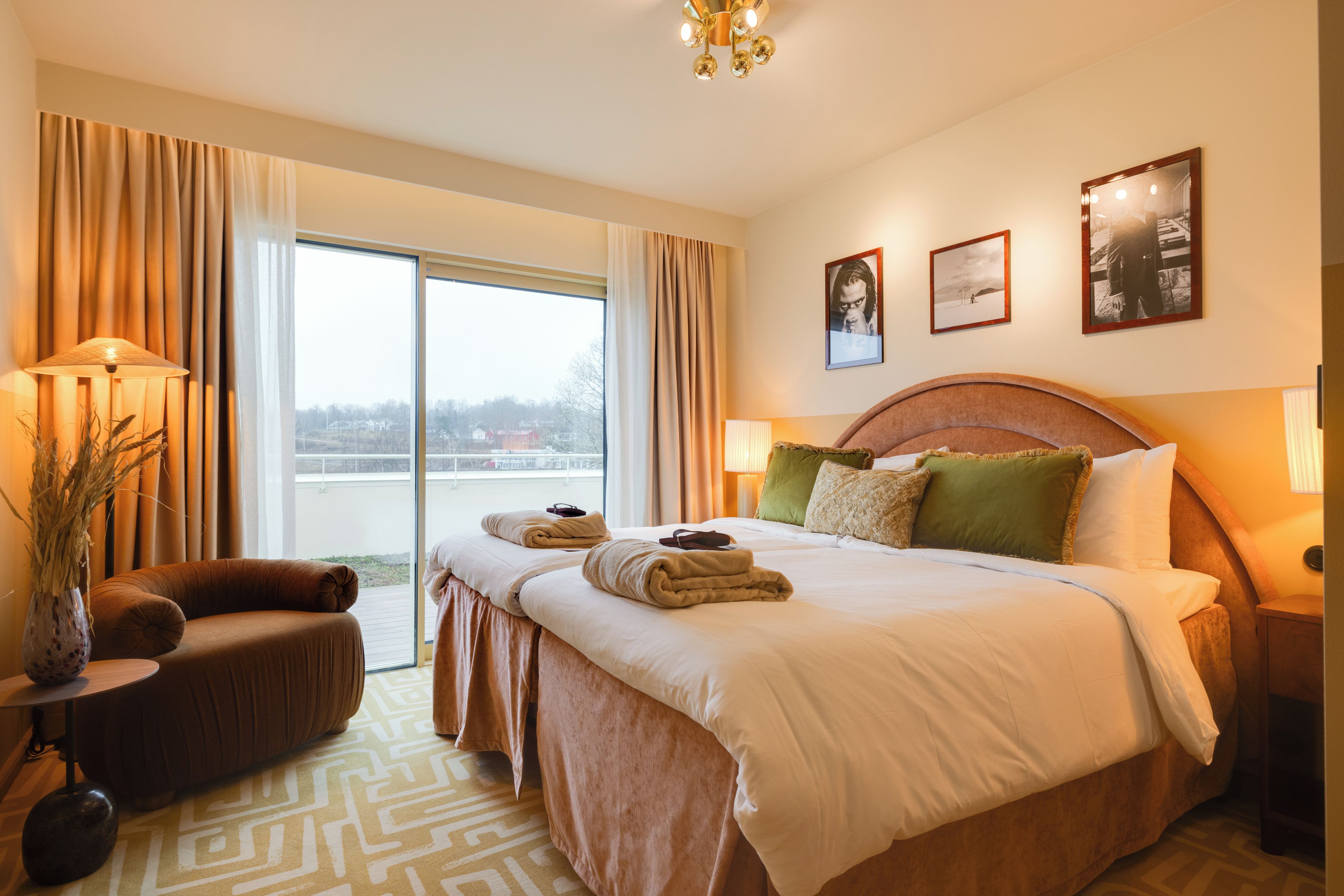 Ett hotellrum med en stor säng, en sittgrupp med en fåtölj, utsikt genom fönstret och en kristallkrona.