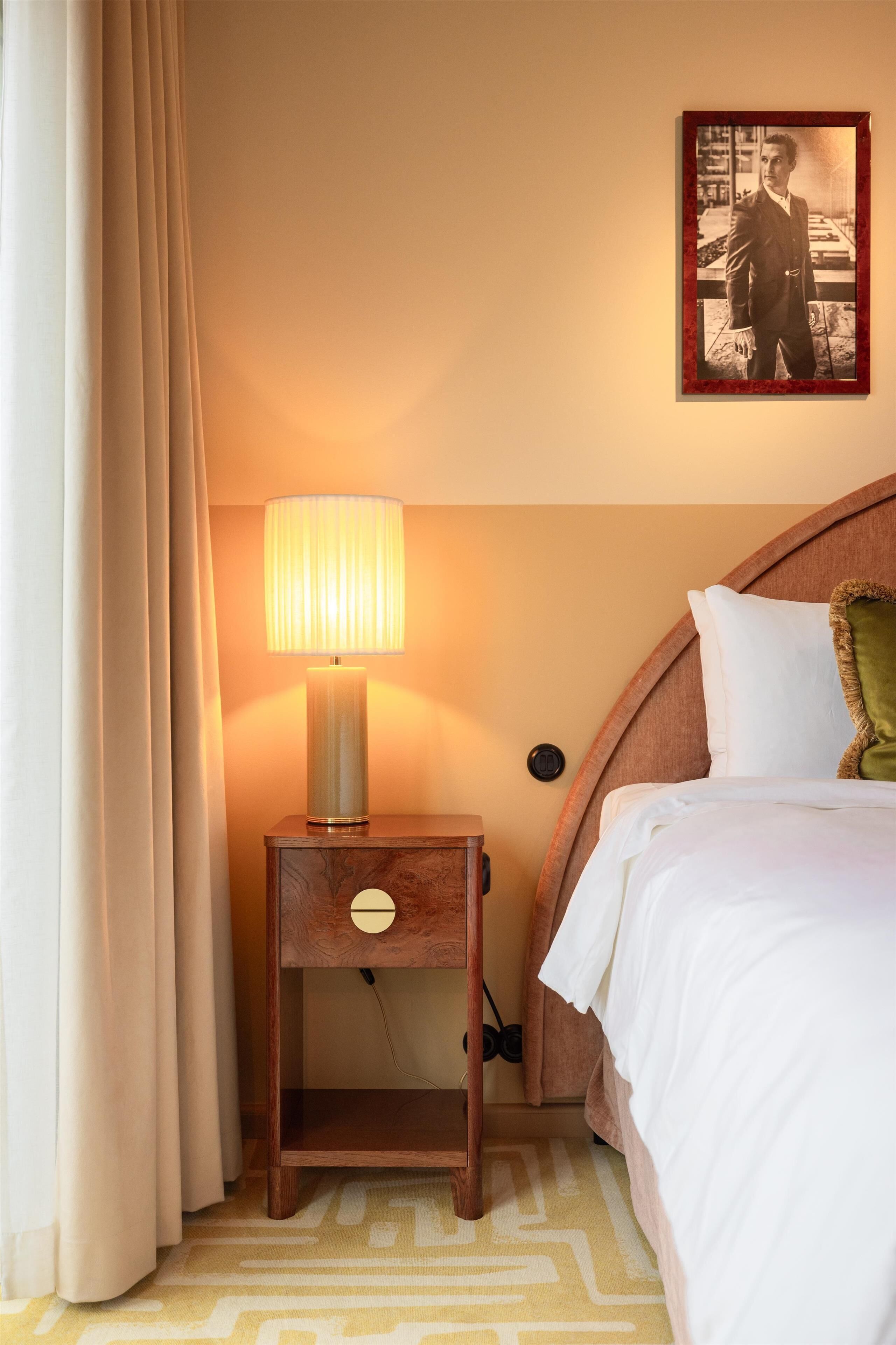 Habitación de hotel con cama, mesita de noche con lámpara, cuadro en la pared y alfombra estampada.