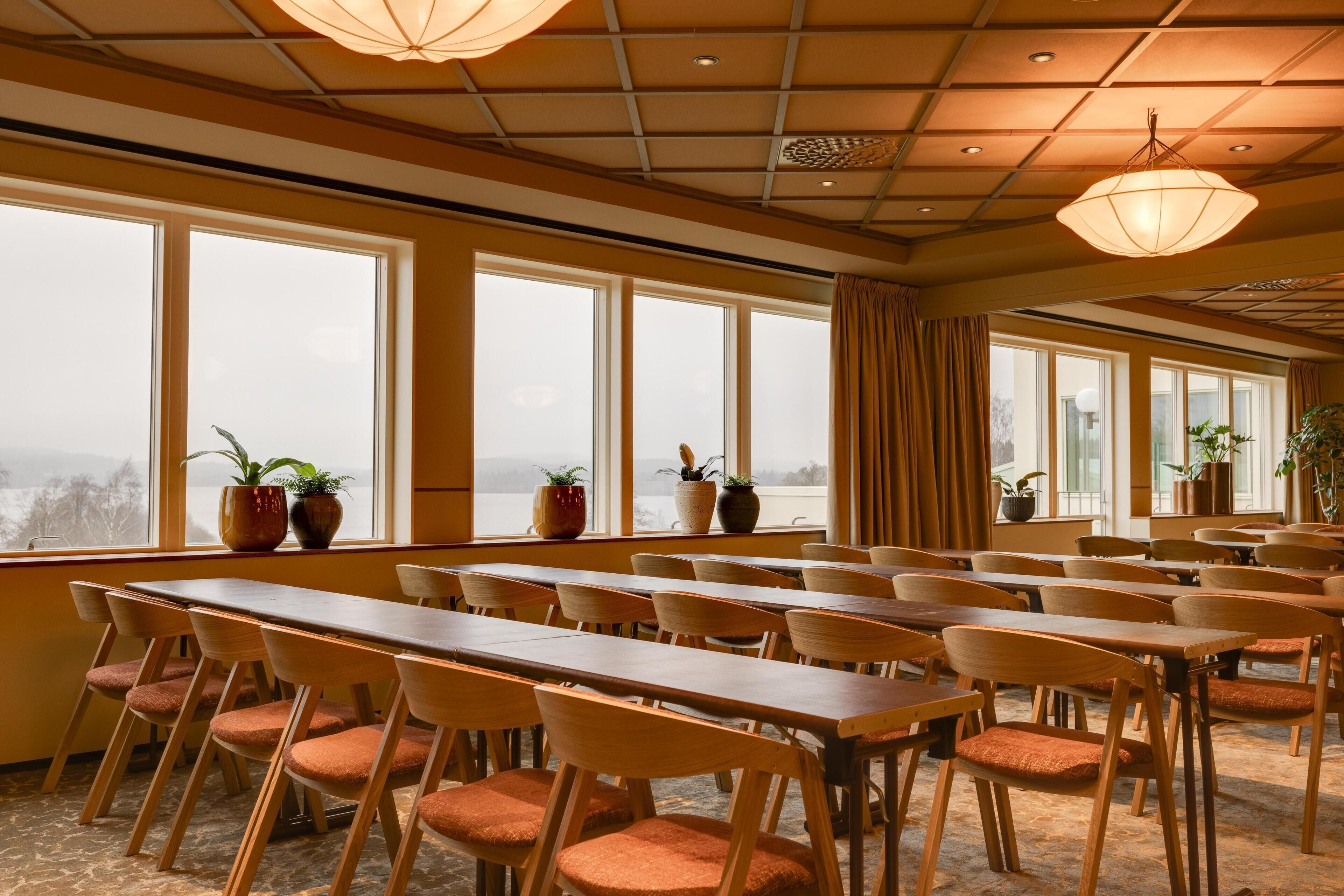 A conference room with long tables and chairs, large windows with curtains, and potted plants.