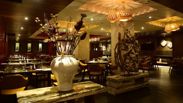 A restaurant with multiple tables and chairs, decorative vases, and hanging light fixtures. - Lemon Tree Hotel Siliguri (Jalpaiguri)