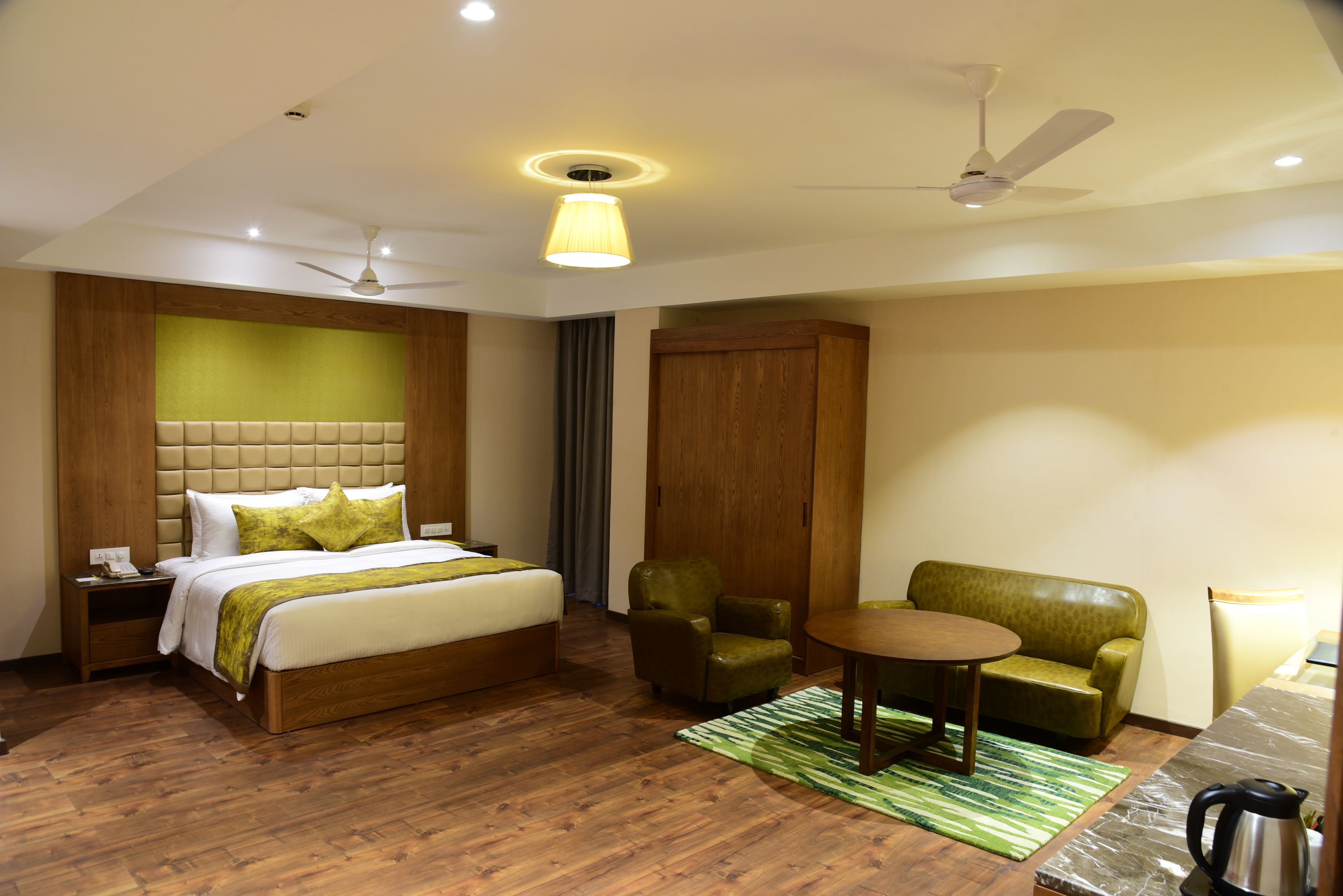 Quarto de hotel com cama, poltrona verde, guarda-roupa de madeira, mesa redonda e ventoinha no teto.
