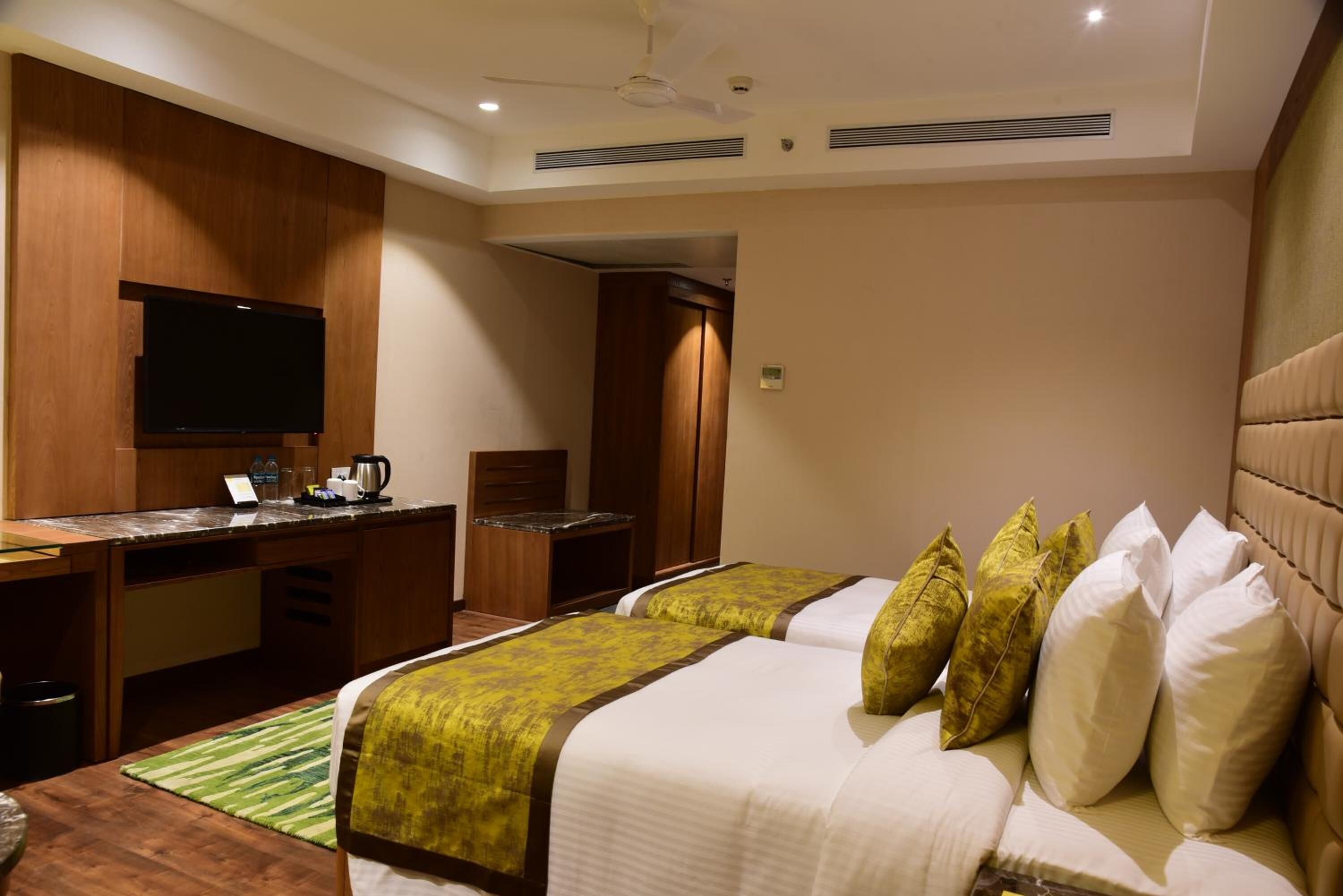 A hotel room with two beds, a flat-screen TV mounted on the wall, a desk with a coffee maker, and a ceiling fan.