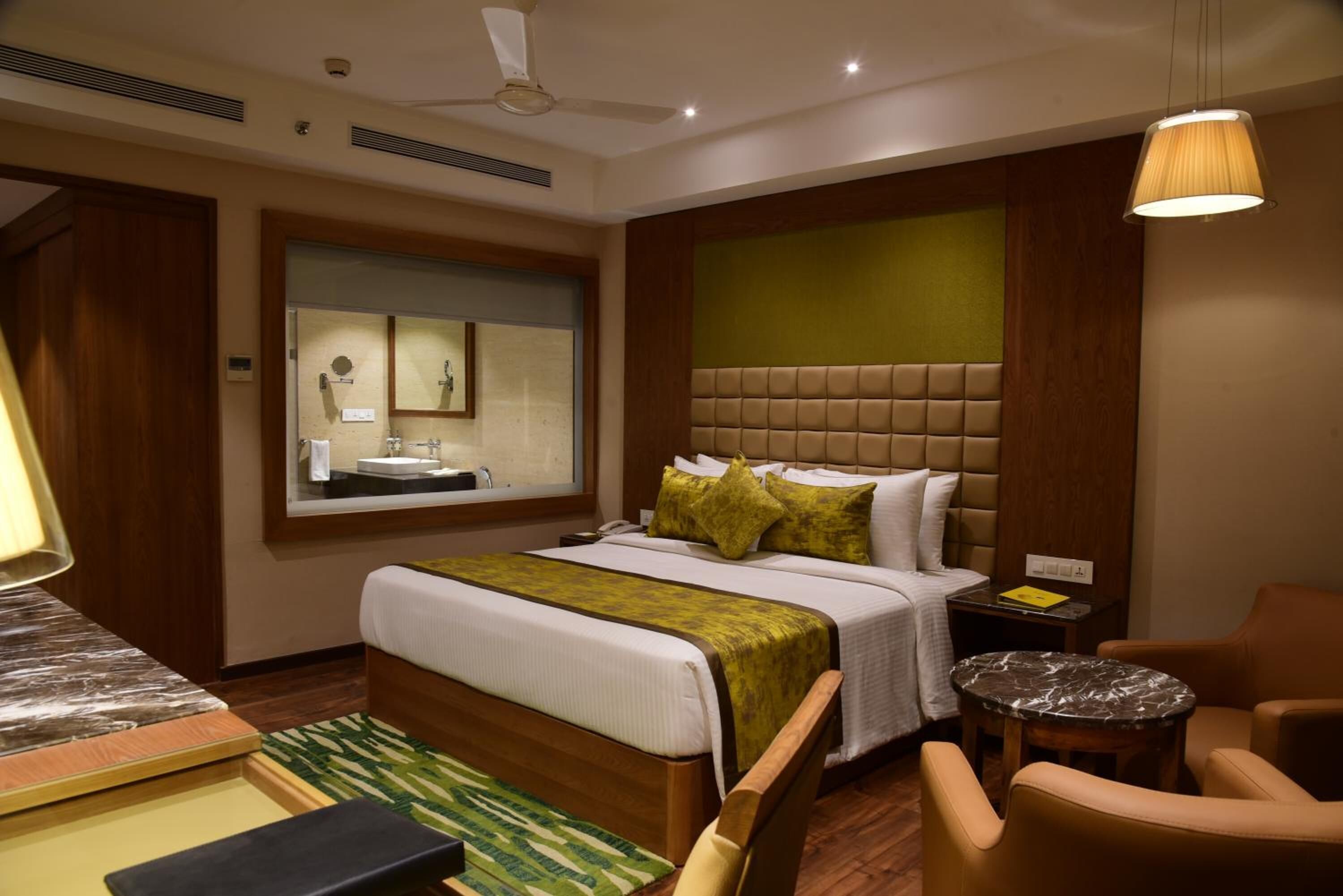 A hotel room with a large bed, a bedside table, a chair, a small round table, a ceiling fan, and a window with a view of the bathroom.