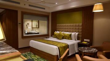A hotel room with a large bed, a bedside table, a chair, a small round table, a ceiling fan, and a window with a view of the bathroom.