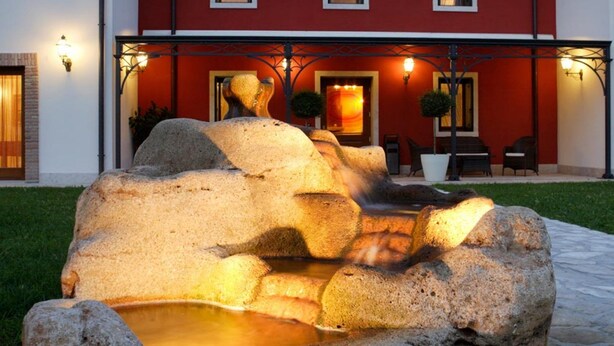 A stone fountain in front of a red building with multiple windows and a door, surrounded by a well-maintained lawn.