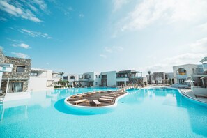 A large swimming pool with wooden loungers and a wooden deck in the center, surrounded by modern buildings with balconies and stone facades. - Ostria Resort & Spa (Ierapetra)