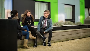 Tre persone sedute su una panchina di fronte a un edificio con pareti esterne verde e nere.