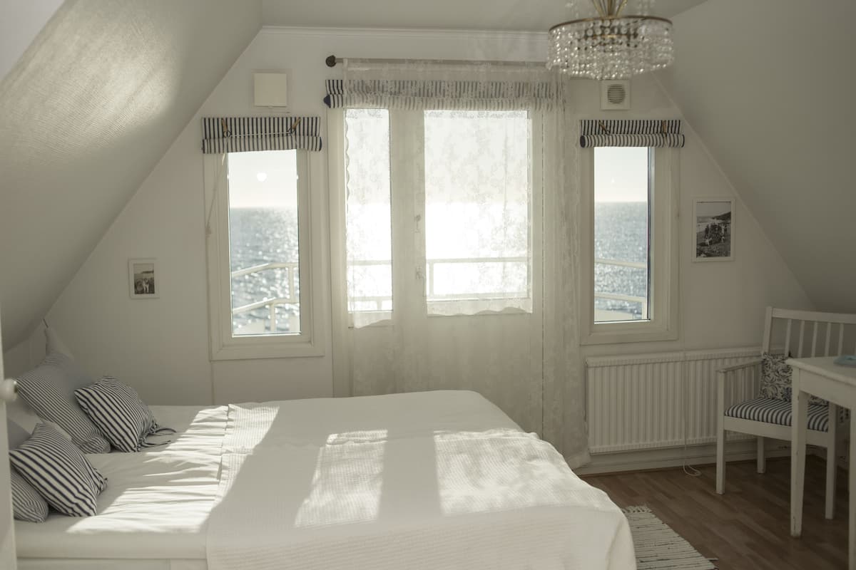 A bedroom with a large bed, two windows with curtains, a radiator, a small table, and a chair.