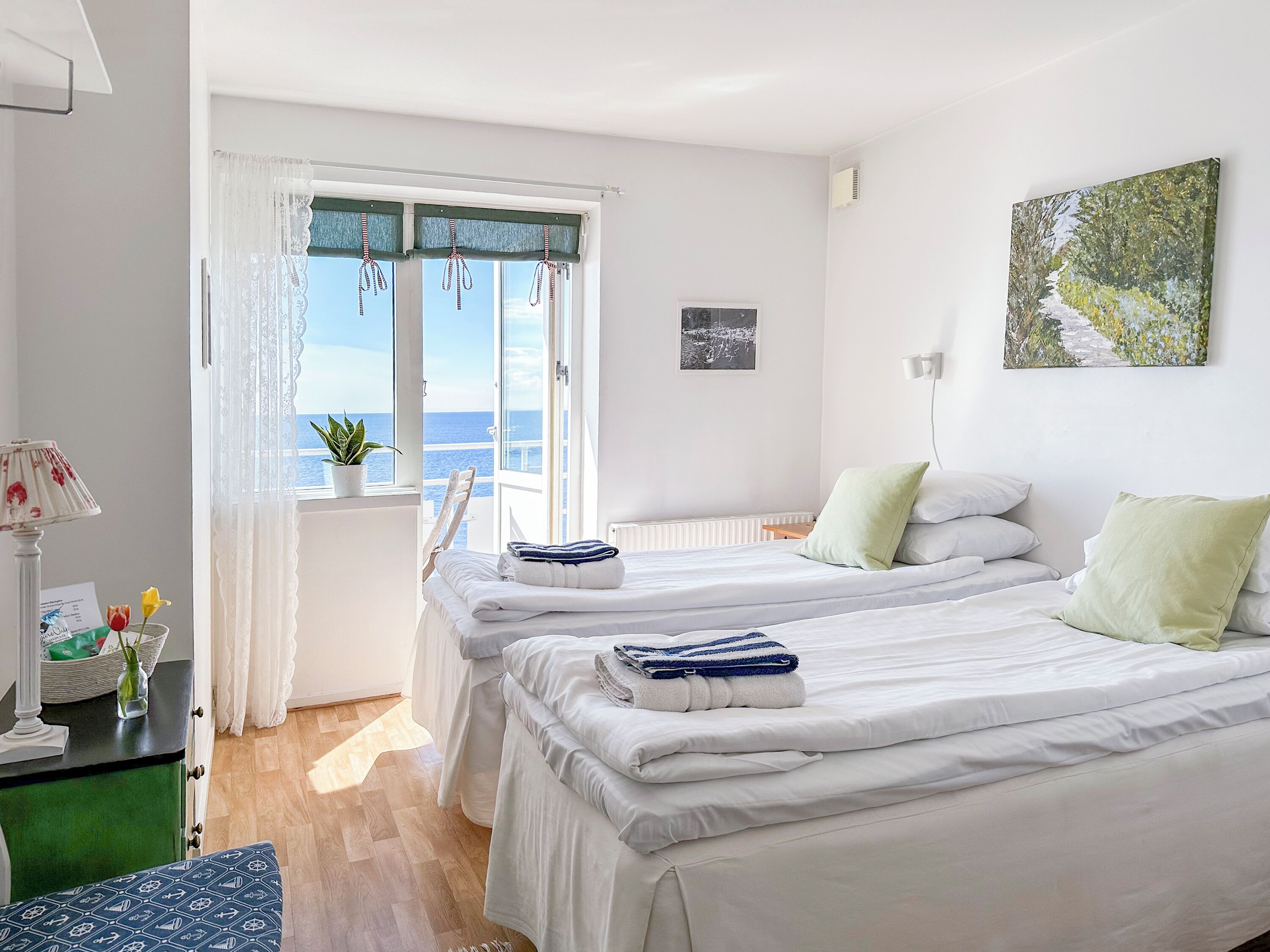A hotel room with two beds, a view of the sea, a potted plant, and a painting on the wall.