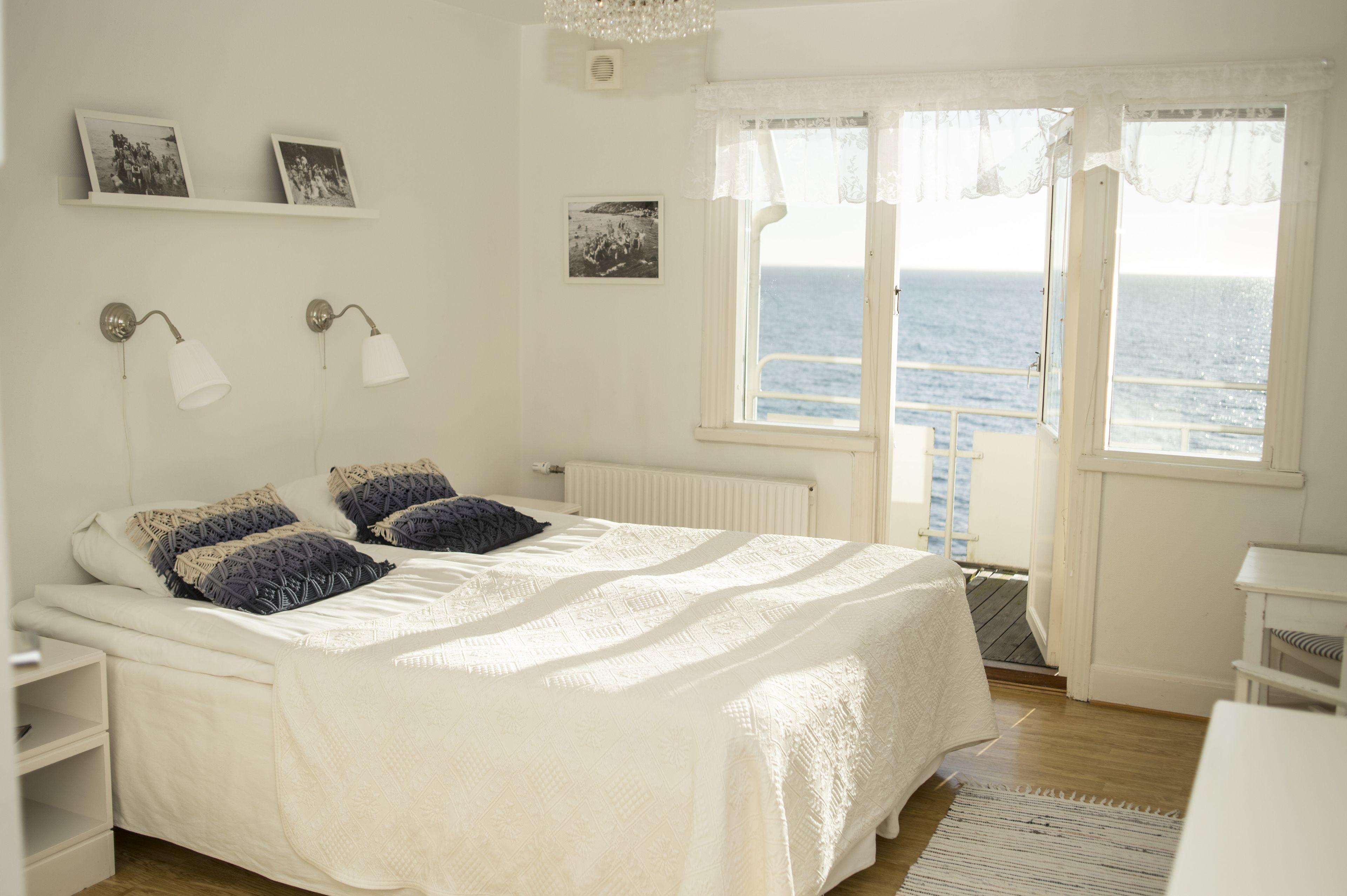 A bedroom with a bed, bedside tables, a nightstand, a window with a view of the sea, and wall-mounted picture frames.