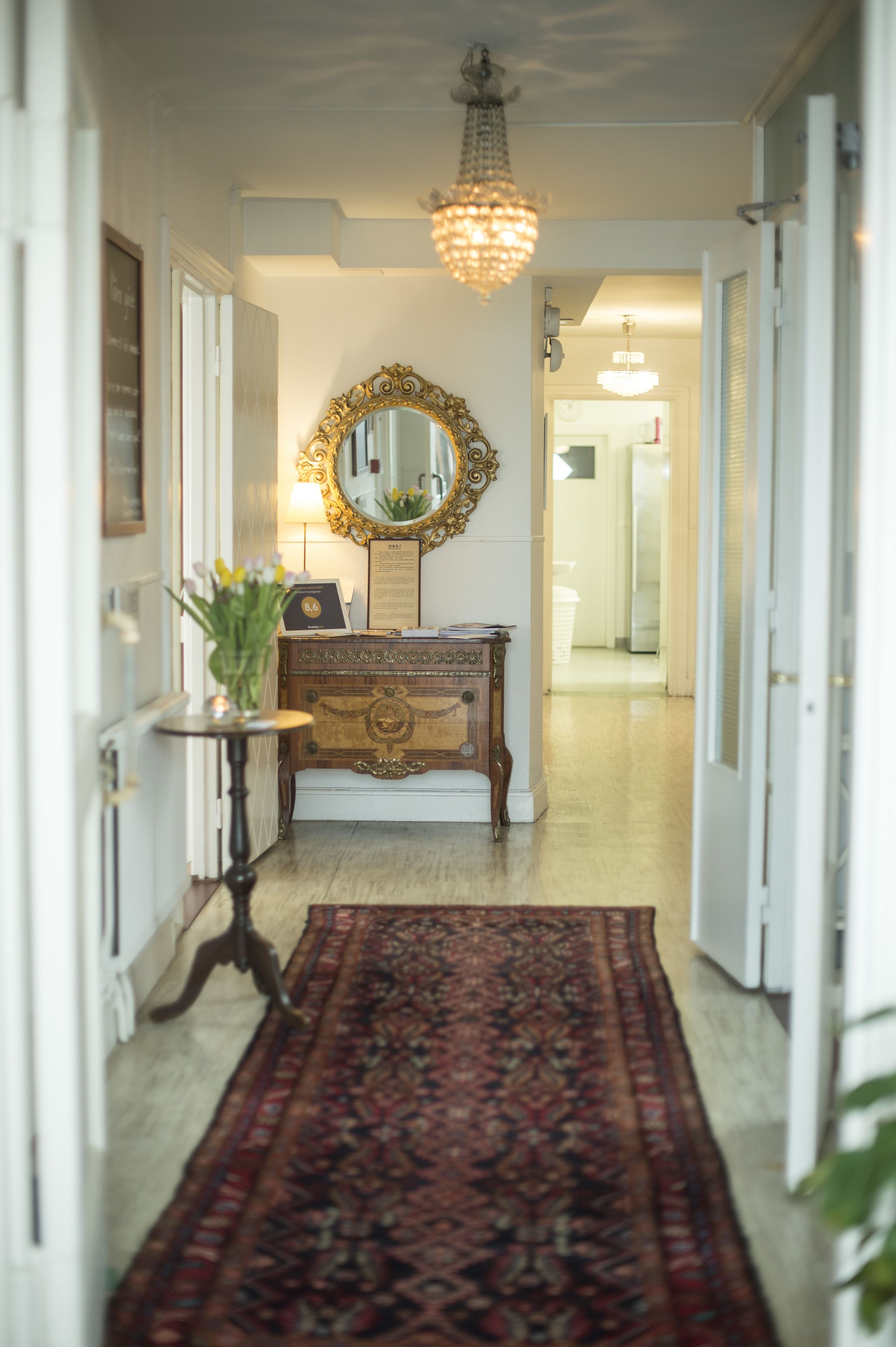 A hallway with a patterned rug, a wooden dresser, a mirror with an ornate frame, and a chandelier.