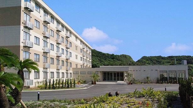 Un hotel de varios pisos con una entrada central, unidades de aire acondicionado en cada balcón y un patio ajardinado con plantas y una palmera.