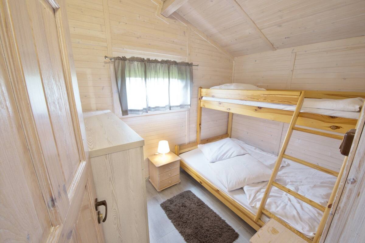 Una habitación de cabaña de madera con literas, una mesita de noche con lámpara y una ventana con cortinas.