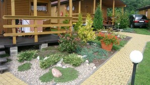 A wooden cabin with a covered porch, a paved pathway, and a garden with various plants and a car parked nearby. - Duet Domki (Postomino)