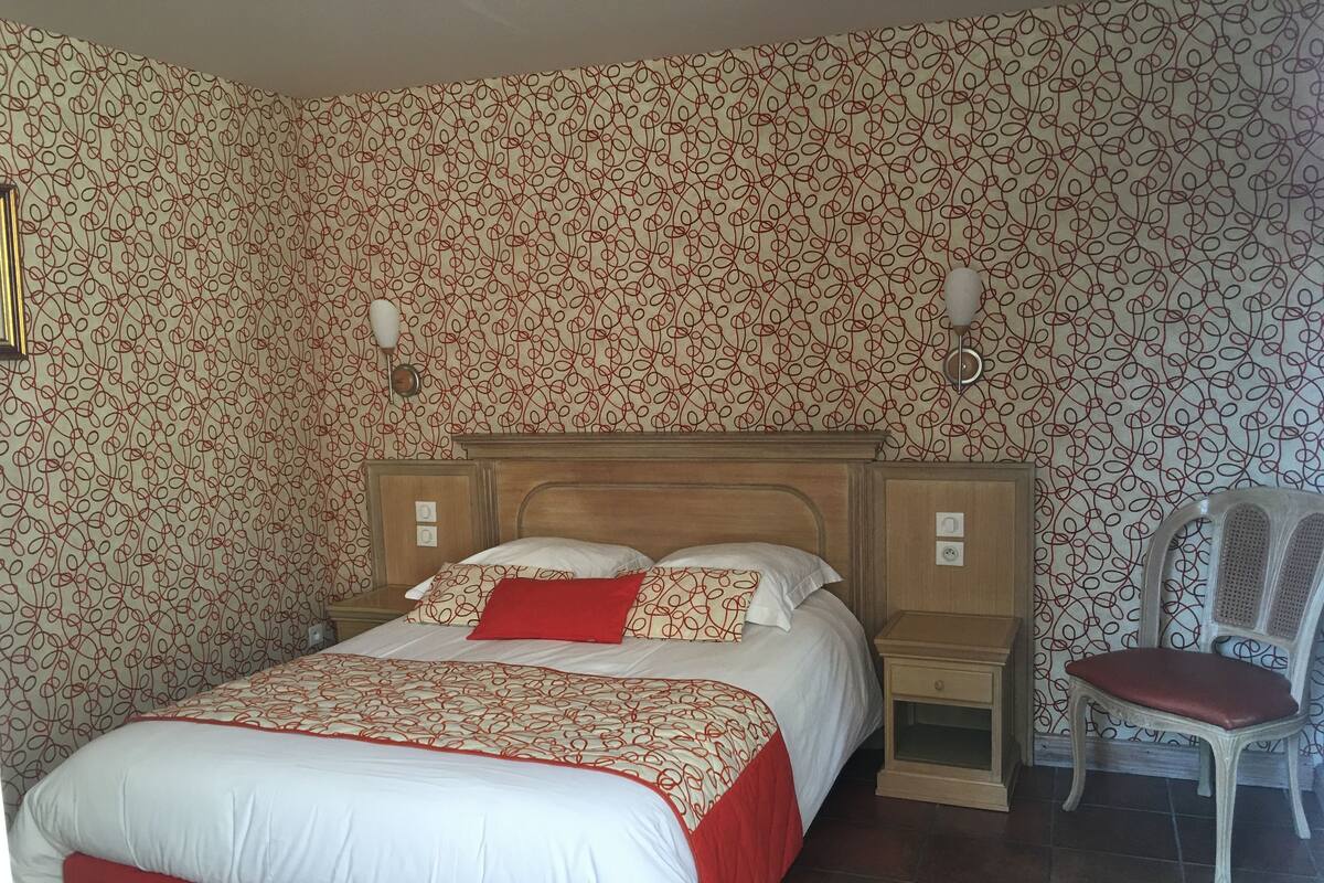 A neatly made bed with a red and white bedspread, two bedside tables, a chair, and patterned wallpaper.