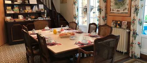 A dining area with a wooden table set for breakfast, surrounded by wicker chairs. A staircase with a wooden railing is visible in the background.