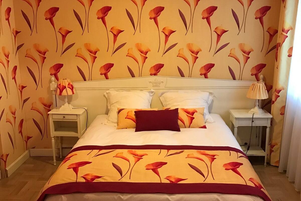 A bedroom with a bed, two bedside tables, and a nightstand. The room has floral wallpaper and a wooden floor.