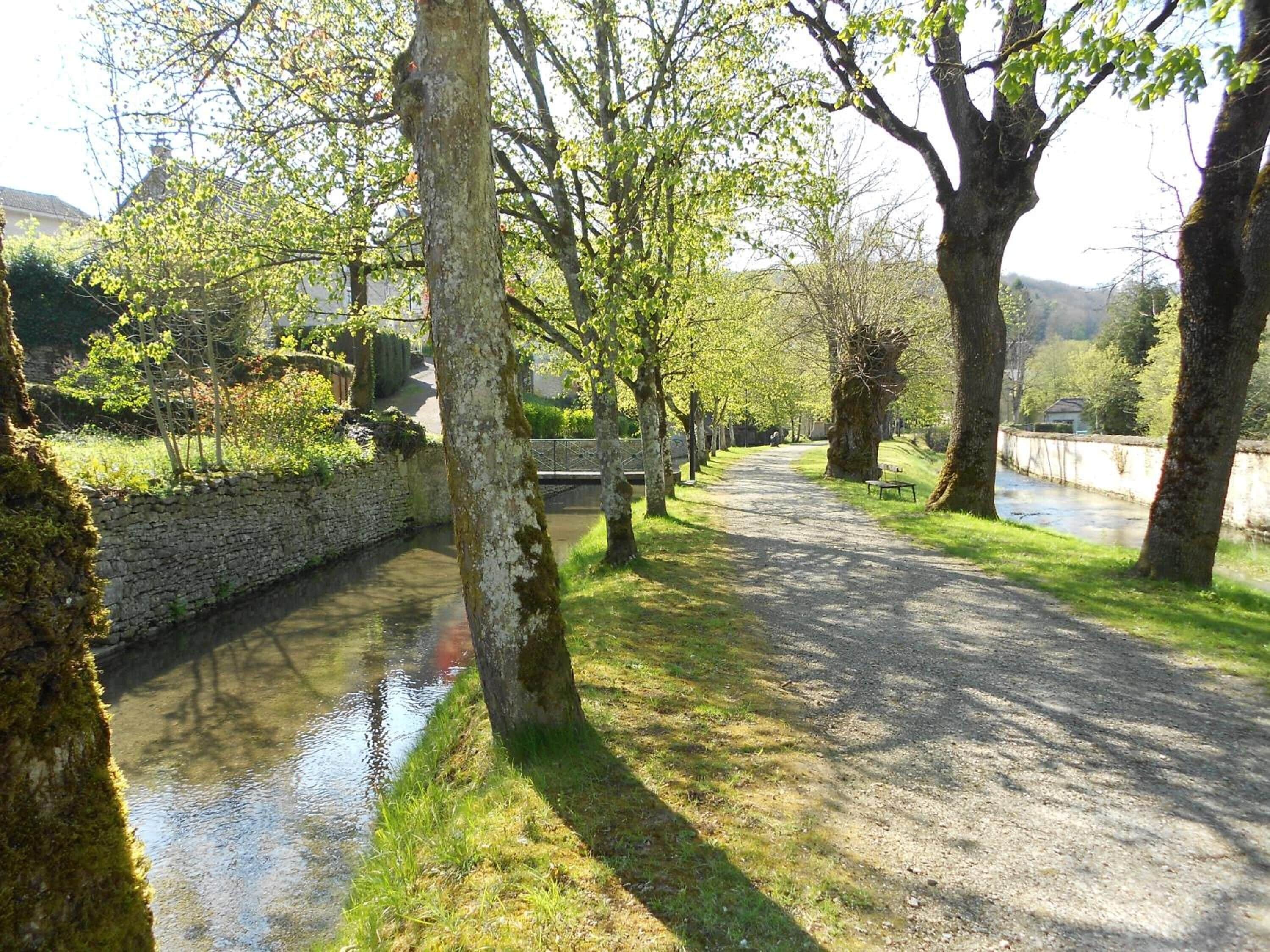 Un sentier paisible le long de la rivière, bordé de murs de pierre et d’arbres, qui mène à un pont.