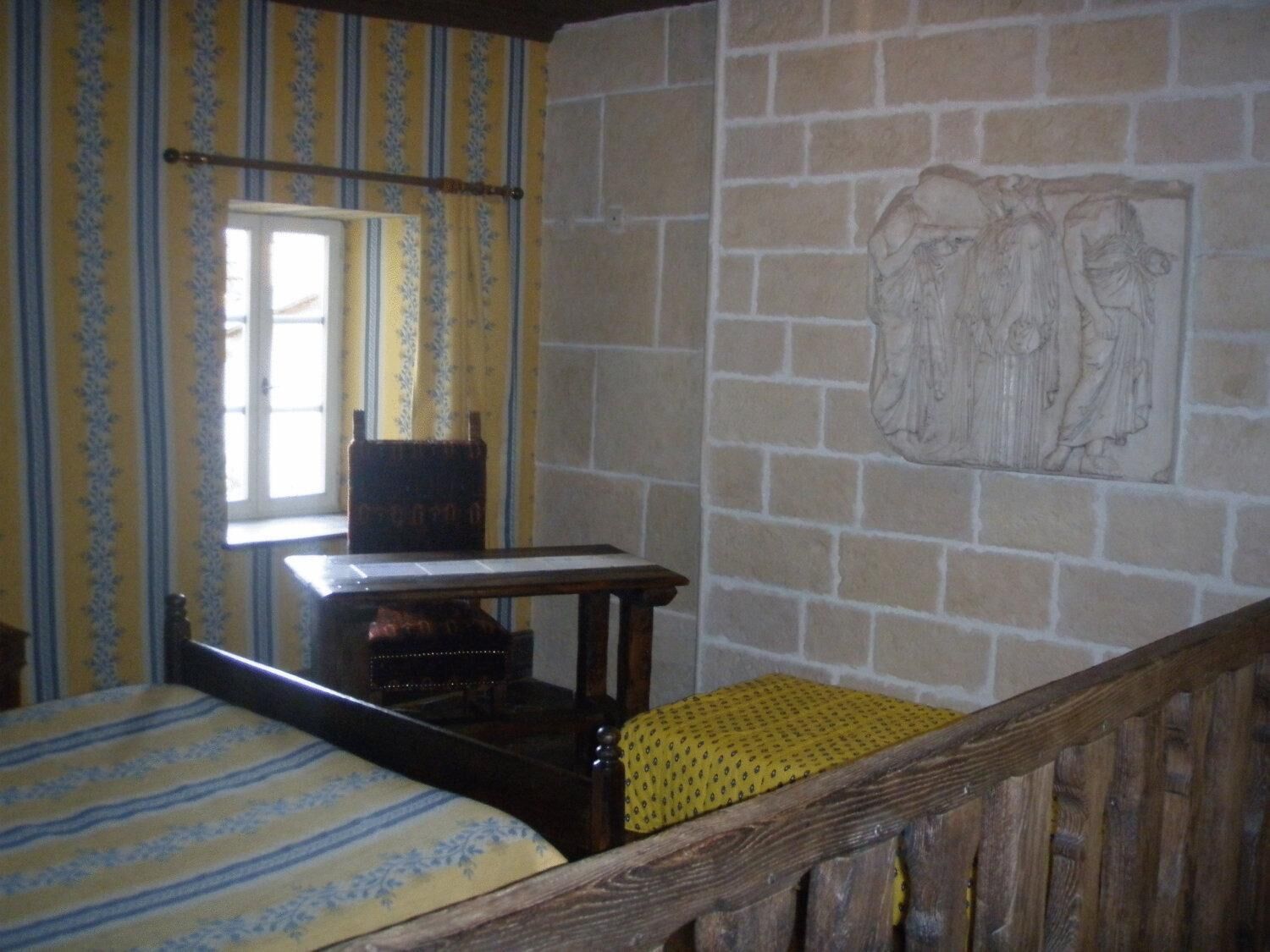 Una stanza con un letto, una sedia di legno, un tavolino e una parete in pietra con un rilievo scolpito.