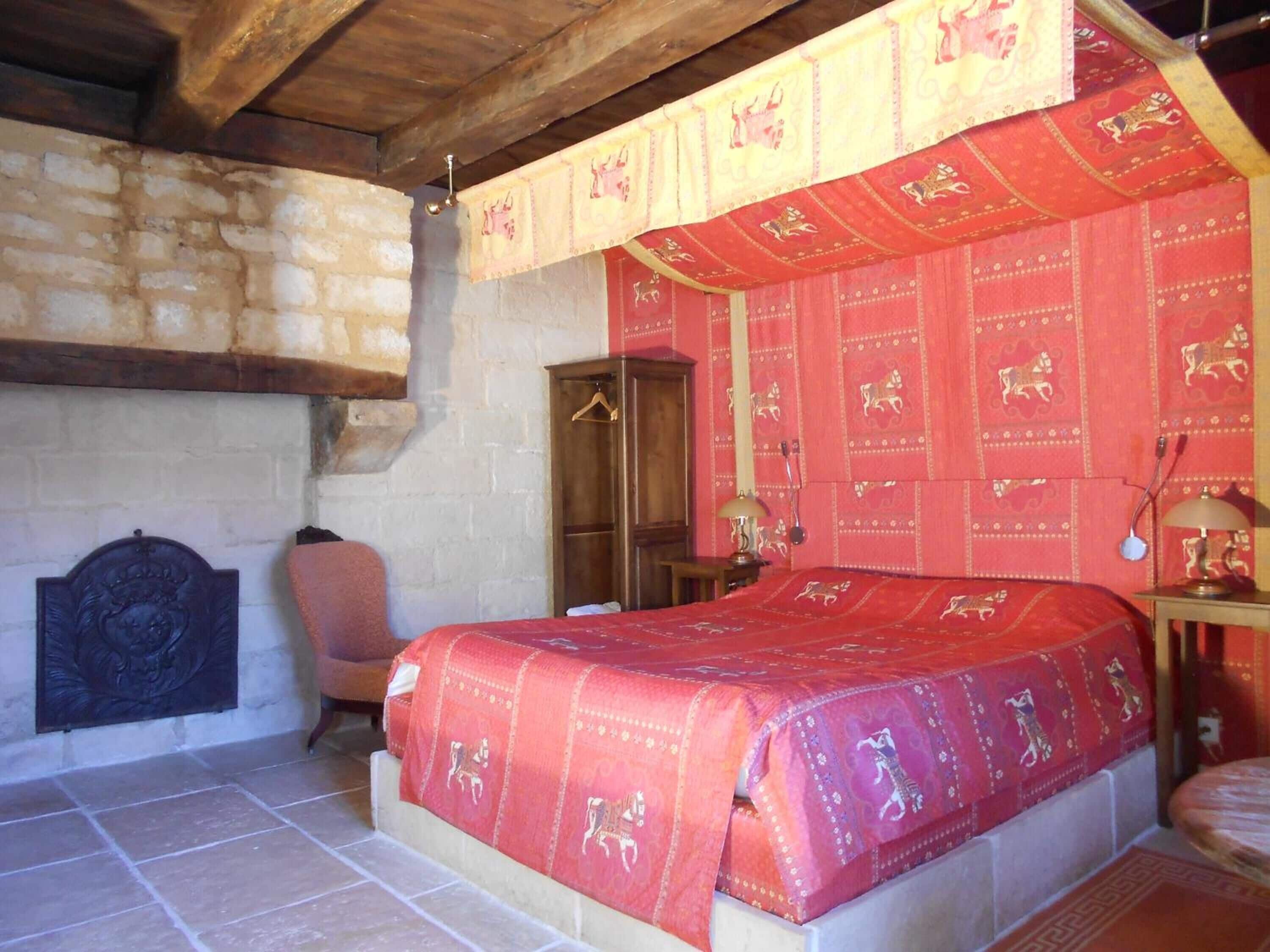 Ein Schlafzimmer mit einem Bett, das mit einer rot gemusterten Tagesdecke bezogen ist, einem Holzschrank, einem Stuhl und einem dekorativen Kopfteil.