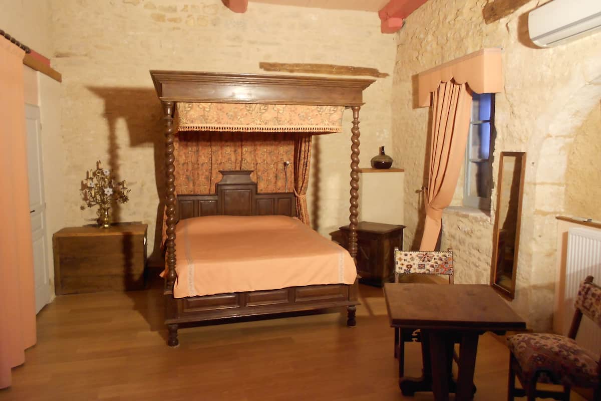 A traditional four-poster bed with a floral bedspread, a wooden nightstand with a vase, and a chair with a patterned cushion.
