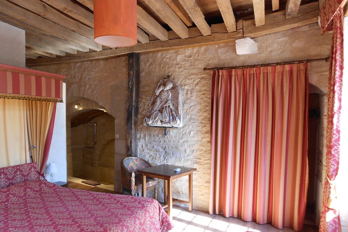 A rustic room with a stone wall, a bed with a patterned bedspread, a wooden table, and a chair.