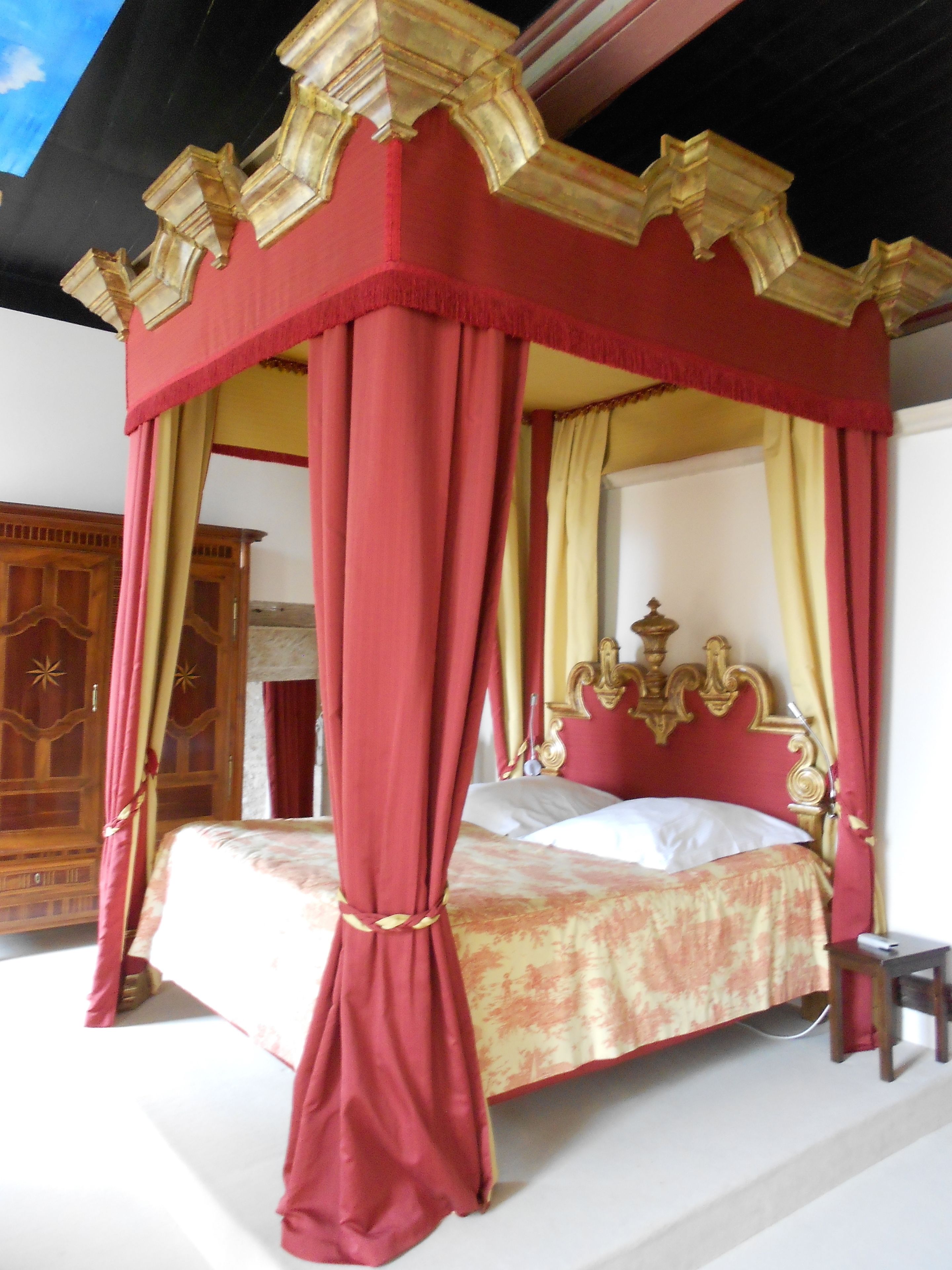 Una cama con dosel adornada con detalles en rojo y dorado, un armario de madera y una mesita de noche.