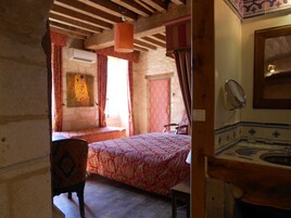 Una habitación con cama, una silla, techo de madera, ventana con cortinas y lavamanos con azulejos.
